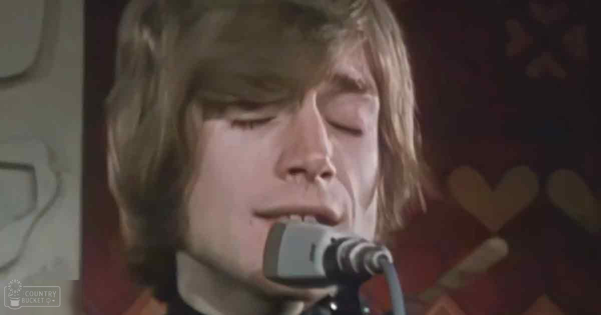A young adult male singer wearing a 1970s-style shirt or jacket with a high collar and long sleeves, holding a vintage microphone during a musical performance in an indoor venue with warm lighting and patterned curtains.
