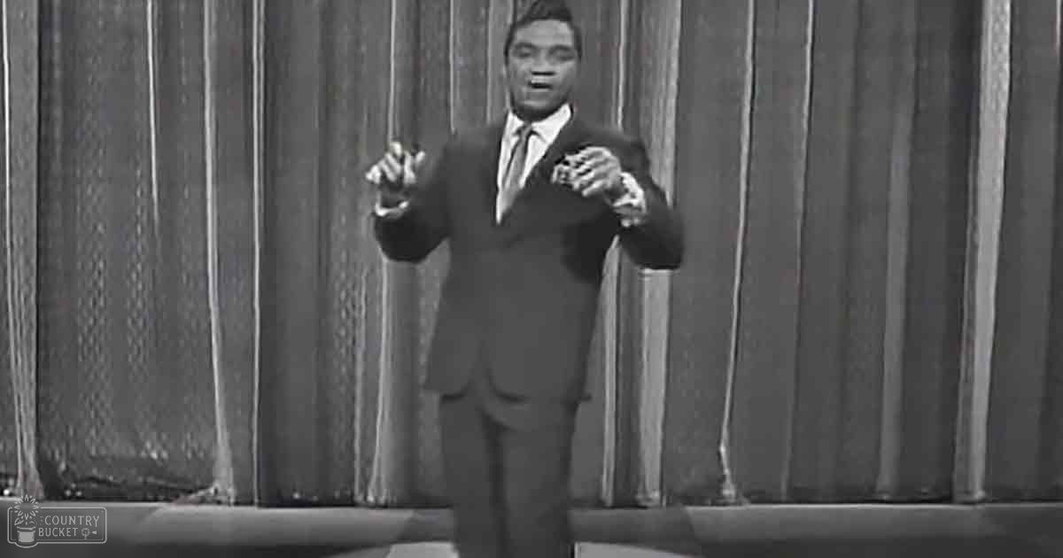 Adult male performer in a dark suit jacket with notched lapel, white dress shirt and dark tie, standing against stage curtains.