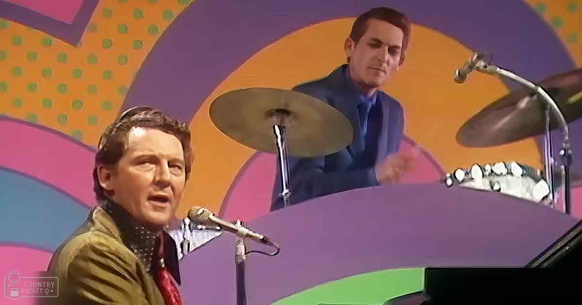 A male adult singer wearing a brown jacket over a white shirt with small dots and playing a piano, accompanied by another adult male drummer in the background performing on stage under vibrant abstract patterns.