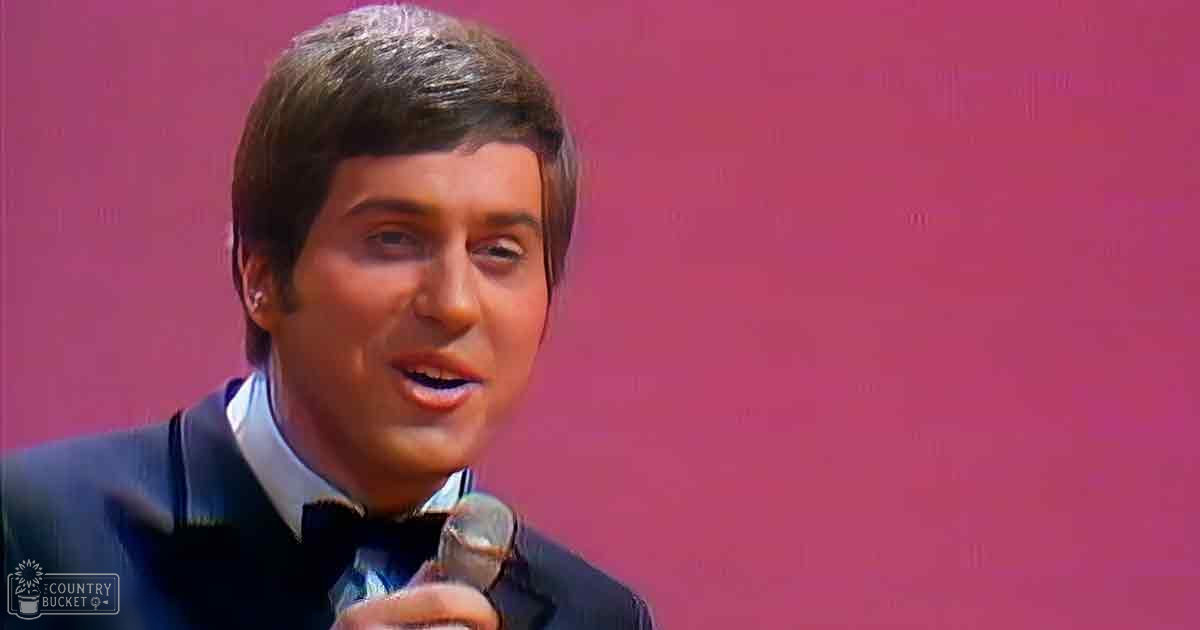 A young adult male in a tailored dark blue suit jacket with a bow tie, speaking or singing into a microphone against a solid pink background.