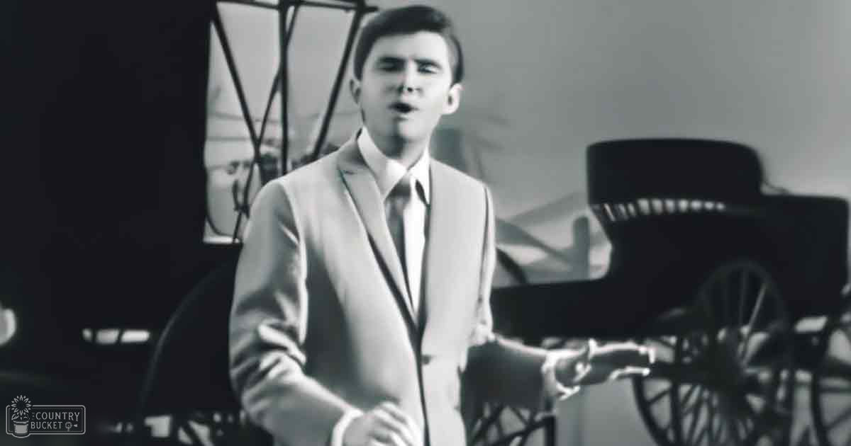 A young adult male wearing a tailored suit jacket and trousers indoors with soft lighting and vintage decor elements.