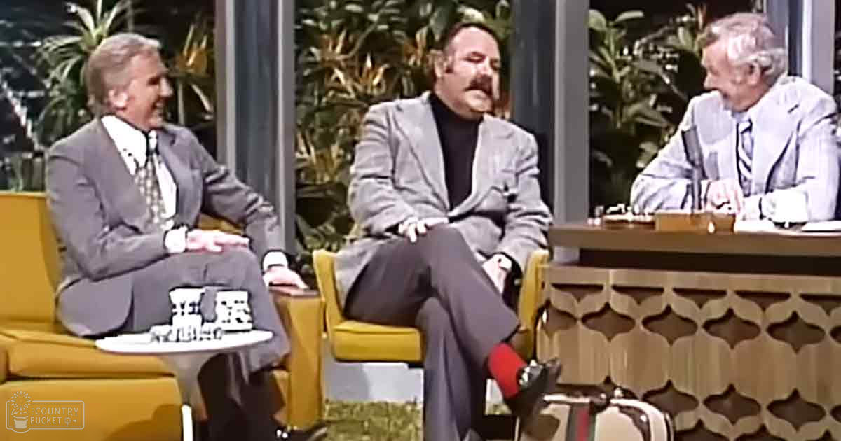 A talk show set with three adult men seated in a studio environment with yellow armchairs, large windows with greenery, and modern furniture from the late 20th century.