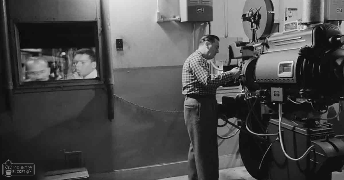 Two adult males dressed for work, standing indoors with a film camera and an indoor set visible through a window, likely in a film studio.