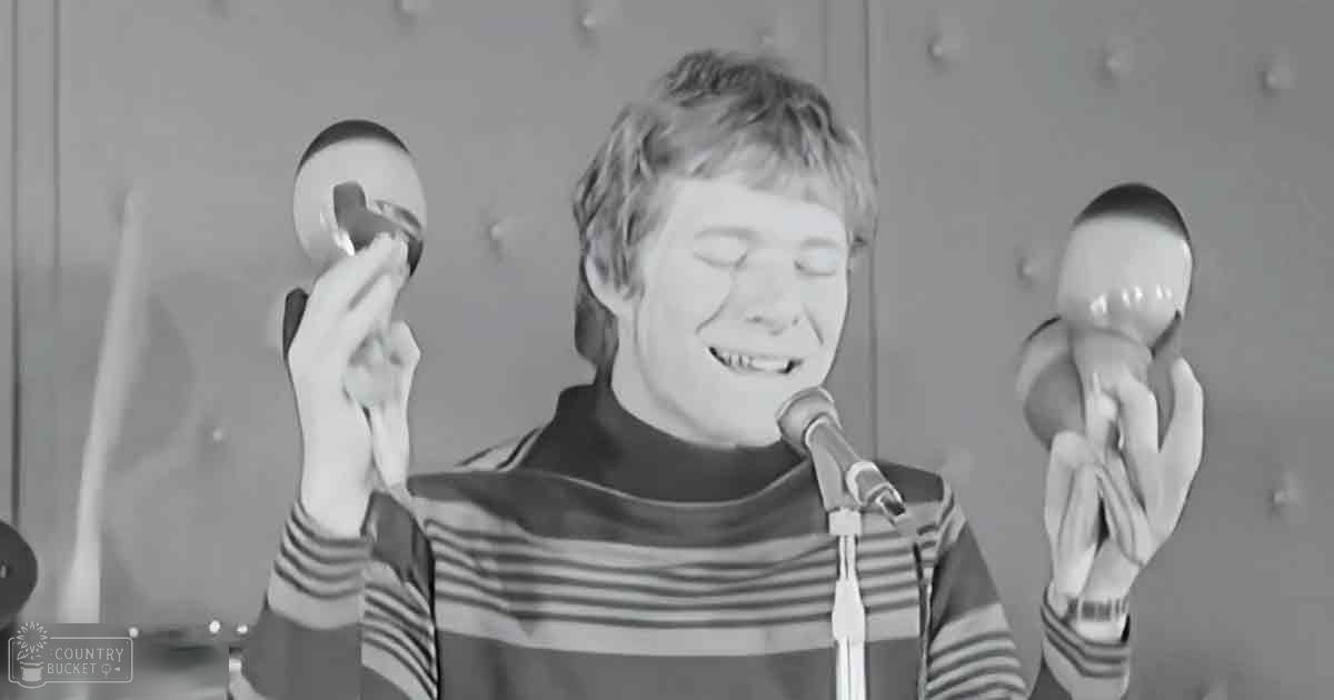 A young adult male in a relaxed, multicolored striped sweater holding maracas stands in front of a microphone stand and textured wall, suggesting a music practice or performance setting.