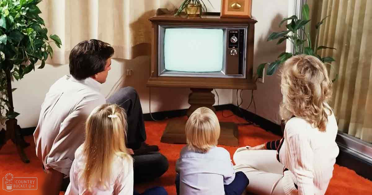 A family of four watching television indoors in a living room with orange carpet and beige curtains.