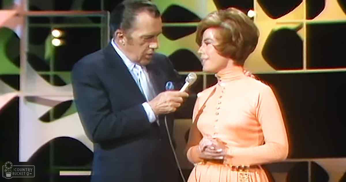 An adult male singer in a dark suit jacket stands on stage with an orange-dressed woman, both under bright artificial lighting against abstract geometric patterns.
