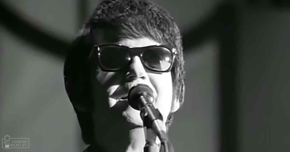 A young adult male in a dark shirt sings into a microphone under dramatic stage lighting, set against an indoor performance backdrop.