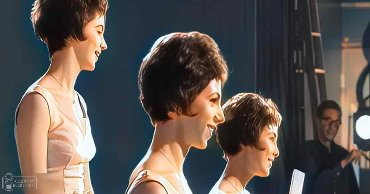 A group of young adults, possibly performing or rehearsing in a dimly lit indoor setting. The individuals are wearing form-fitting white sleeveless tops with thin straps and a gold clasp at the neckline.