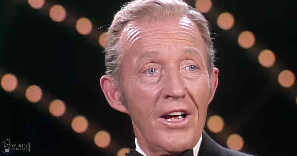 A middle-aged to older adult male in a dark suit jacket at a formal event with blurred stage lighting, suggesting an indoor venue.