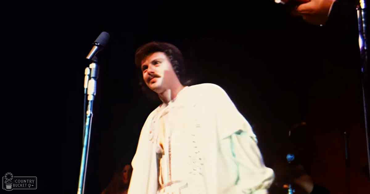 Adult male performer in a white, high-collared shirt with decorative buttons on stage under focused lighting during a live event.