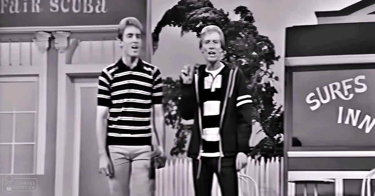 Two young adult males wearing casual clothing stand indoors with a backdrop resembling a beachside town; one wears a striped polo shirt and khaki pants, the other a horizontally striped sweater over a collared shirt with dark pants and a watch. A 'Surf's Inn' sign is visible to their right.