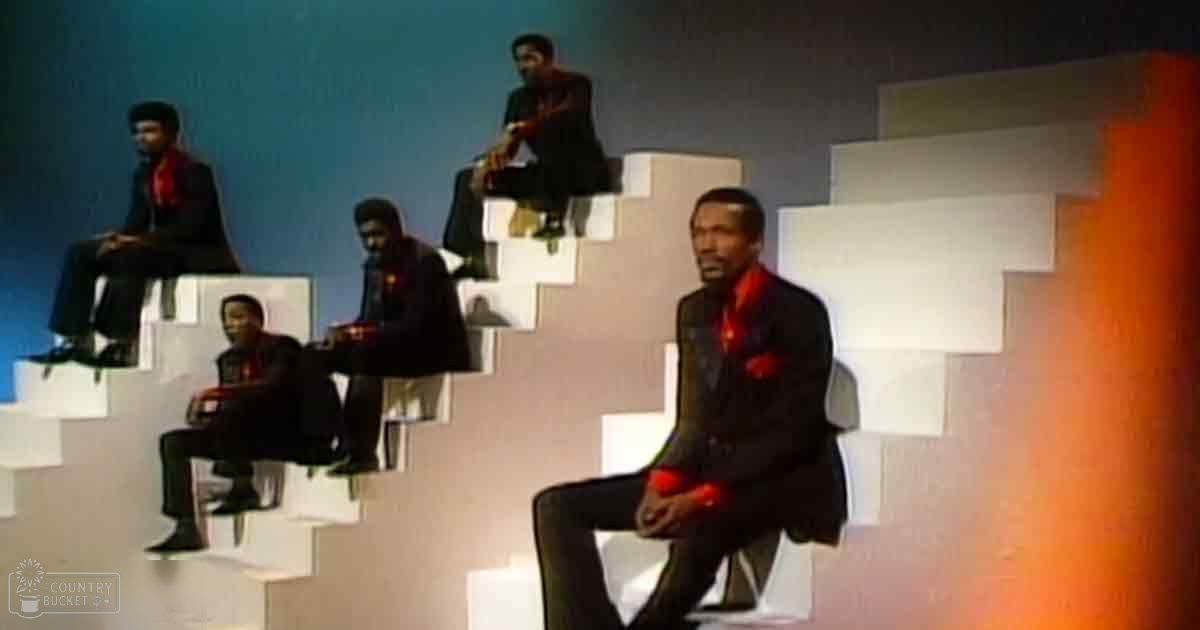 Four adult males in dark suits with a red scarf seated on geometrically shaped structures indoors with muted background colors