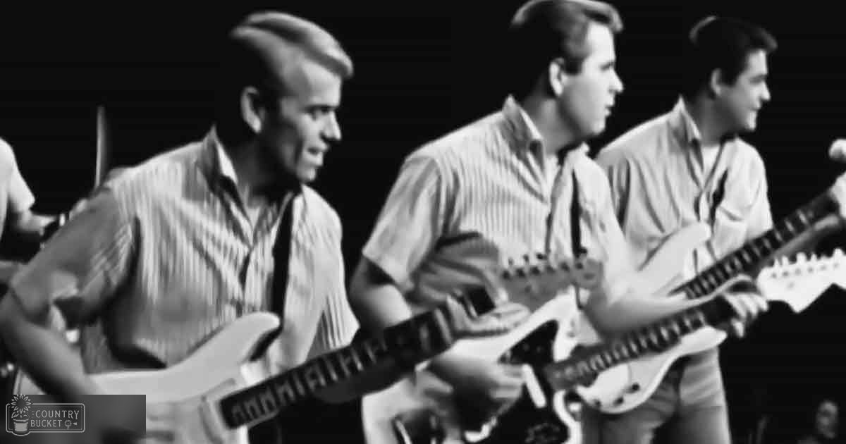 A musical performance scene with three young adult male musicians playing electric guitars on a dimly lit stage, possibly from the mid-20th century.