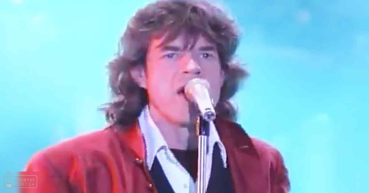 An adult male singer in a red jacket with a zipper at a concert setting, wearing a white shirt and dark undershirt, surrounded by stage lighting and a microphone against a light blue backdrop.