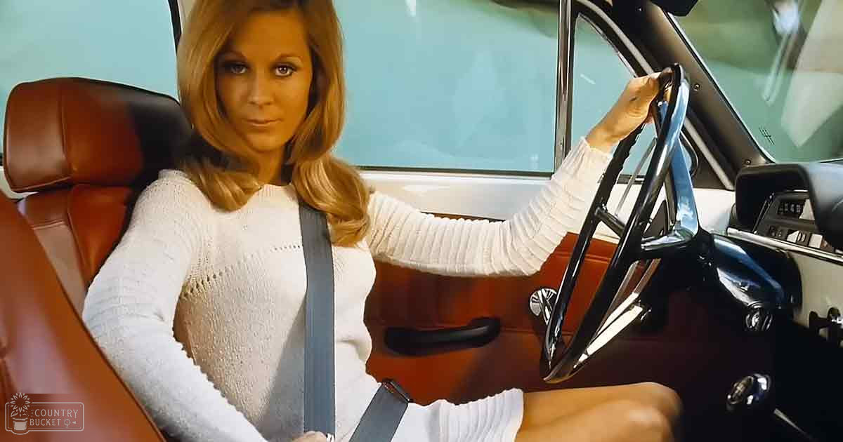 A middle-aged woman wearing a white long-sleeved sweater with ribbed cuffs and neckline, seated inside an older model car with brown leather seats during daytime.