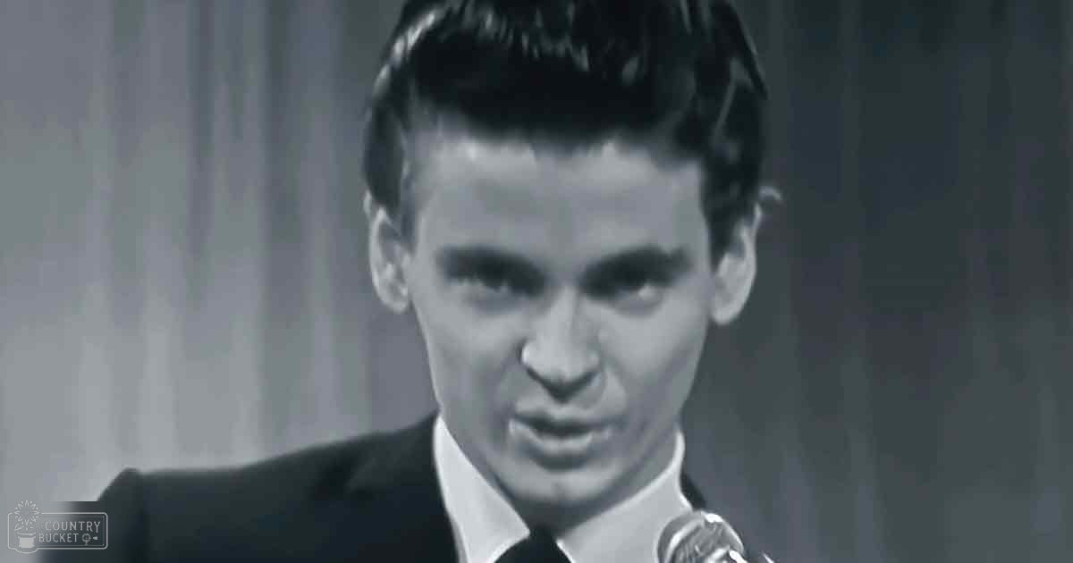 A young adult male in a dark, fitted suit jacket with a tie stands indoors near a microphone, possibly at an event or performance.