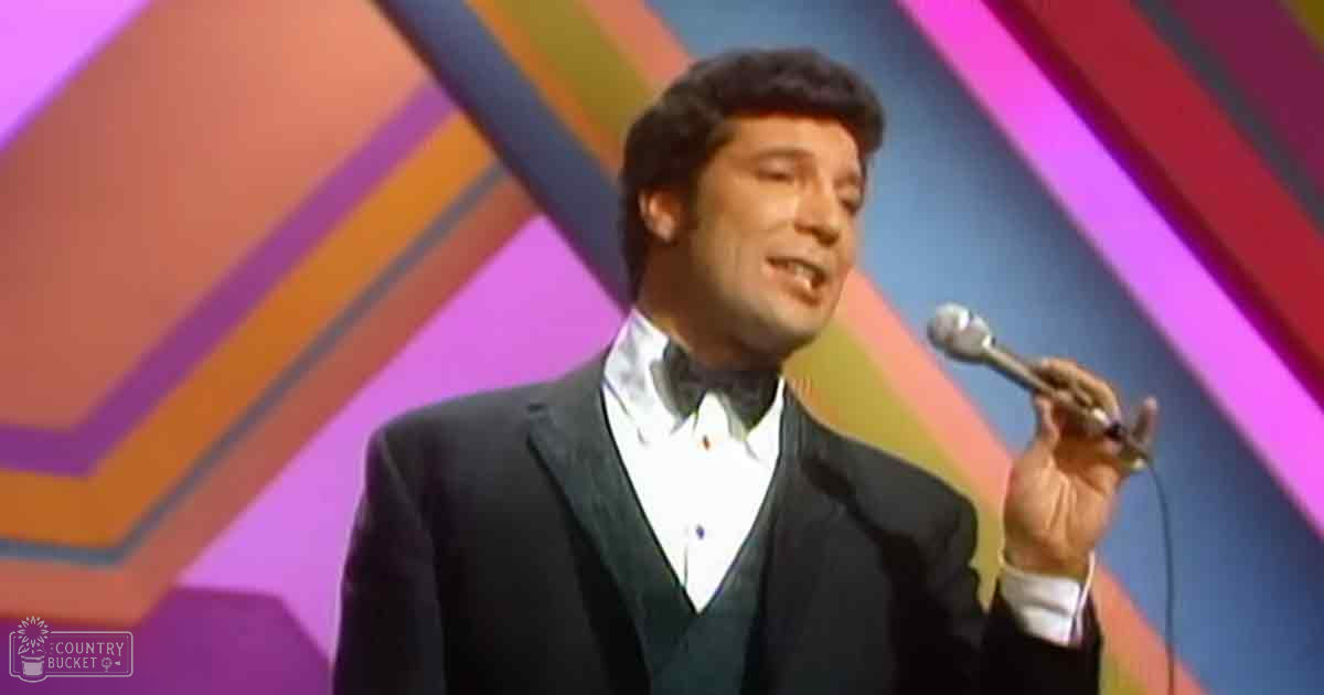 Adult male performer wearing a dark suit with white shirt and bow tie, holding a microphone against a colorful geometric backdrop on an indoor stage.