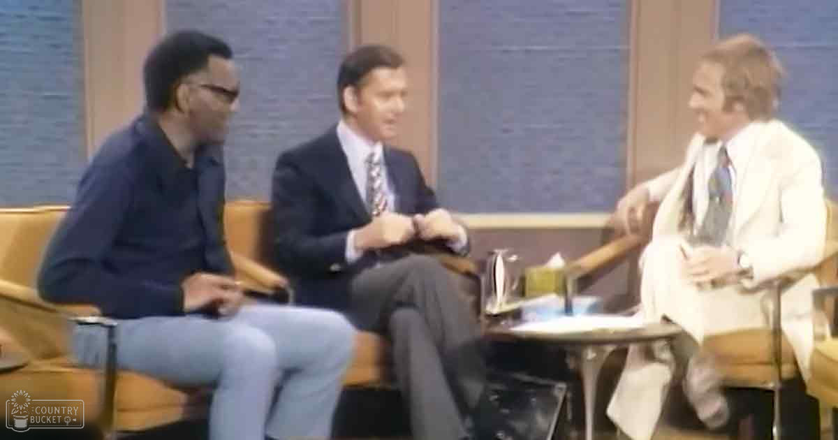 Three adult males seated on upholstered chairs around a small round table in an indoor studio with blue curtains, dressed casually yet professionally for a talk show or interview setting.