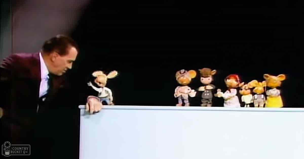 An adult male puppeteer in a dark suit interacts with anthropomorphic puppets resembling animals, set against a dark stage backdrop.