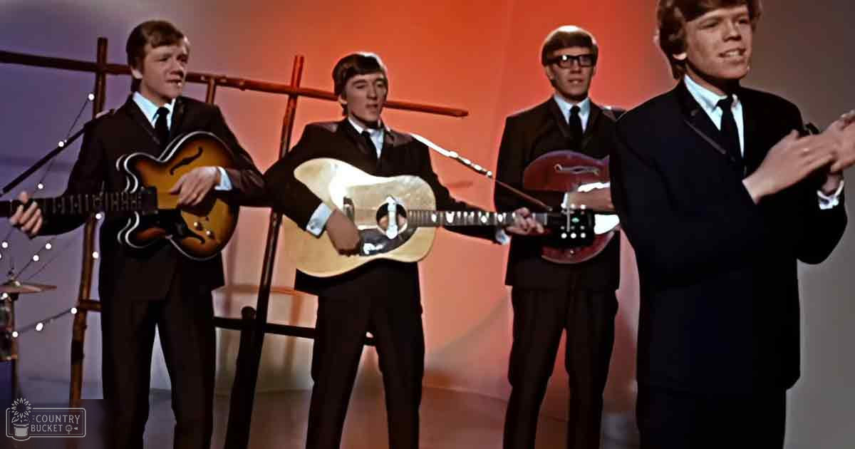 Musical performance group in 1960s attire on stage with electric guitars and drum set.