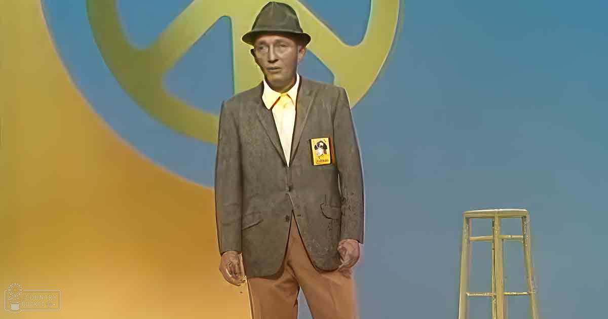 An adult male in a dark gray suit with a yellow tie and fedora, standing against an abstract background resembling a stage or studio.
