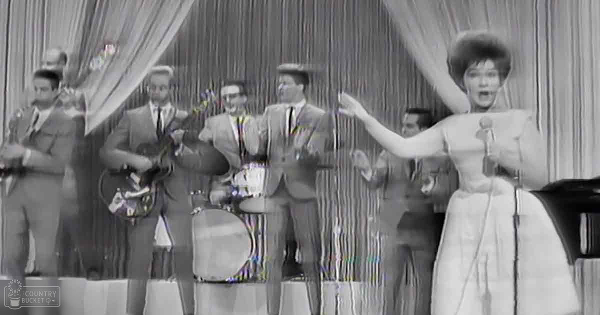 An adult female singer performing in front of a band at an indoor event with mid-20th century attire and instruments, holding a microphone.
