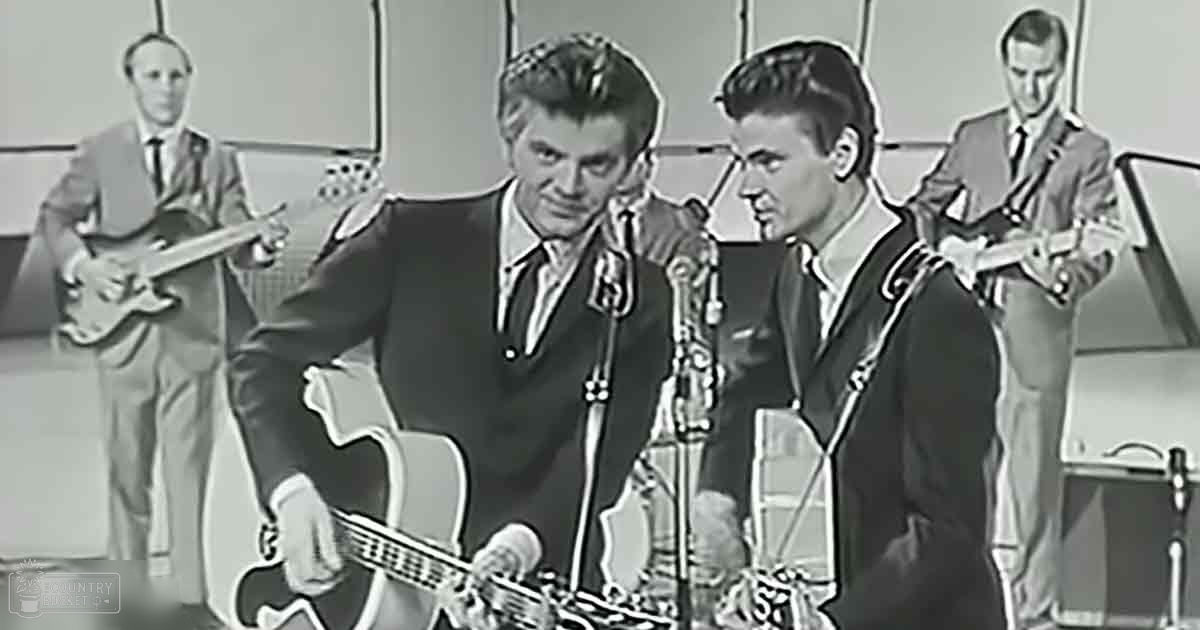 A mid-20th century musical performance scene with four adult male musicians in suits, one holding a guitar over his shoulder against a plain light-colored backdrop.