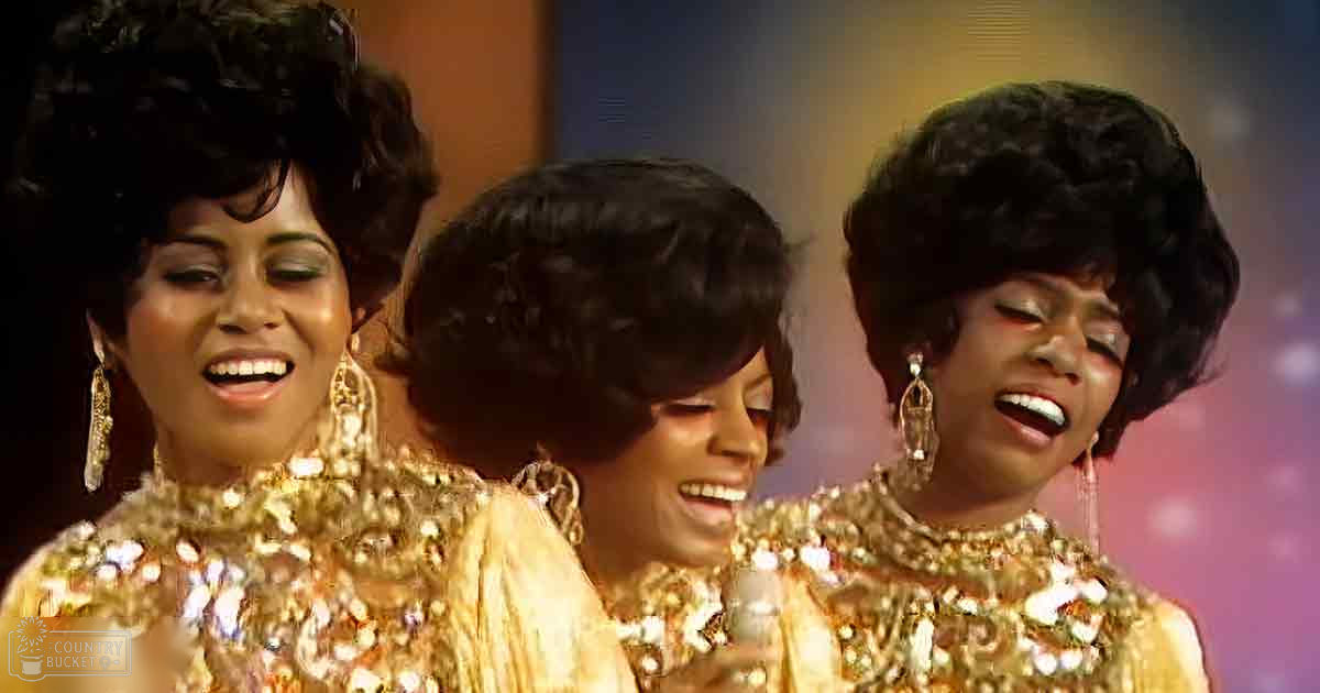 A group performance scene with three adult females wearing matching gold sequined tops, set against a stage backdrop with soft artificial lighting creating a glowing effect.