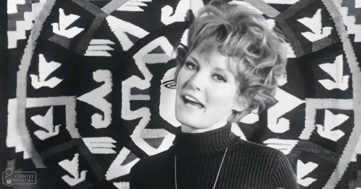 Middle-aged adult female wearing a fitted ribbed turtleneck sweater against an indoor backdrop with abstract black and white geometric shapes, diffused lighting suggesting a studio setting.