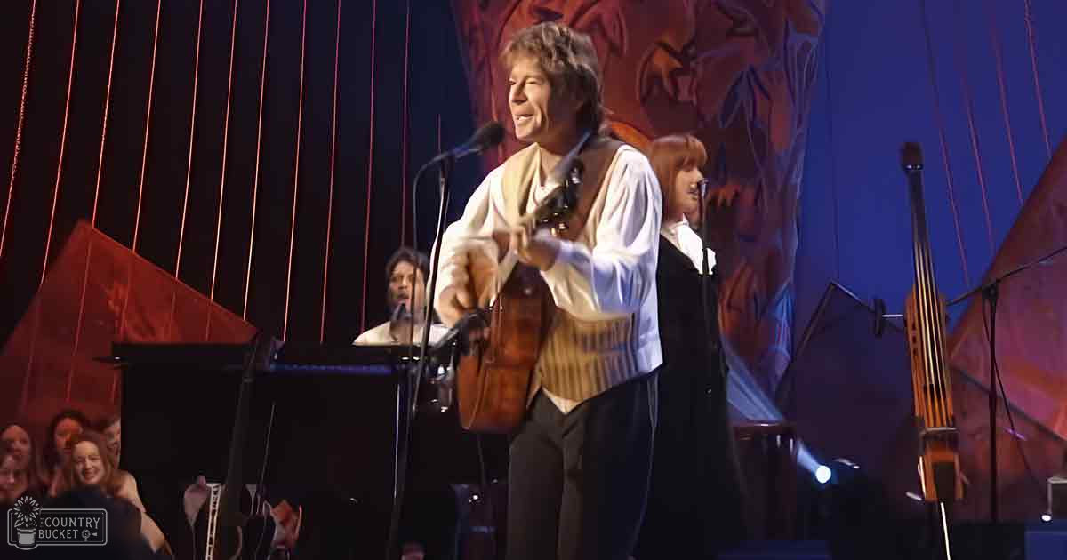 A middle-aged male performer playing guitar, wearing a white long-sleeve shirt and brown vest over dark pants, with an adult female singer in a black outfit to his left and another middle-aged male pianist in dark clothing. The scene is set in a dramatic indoor concert hall or theater with stage lighting focused on the performers.