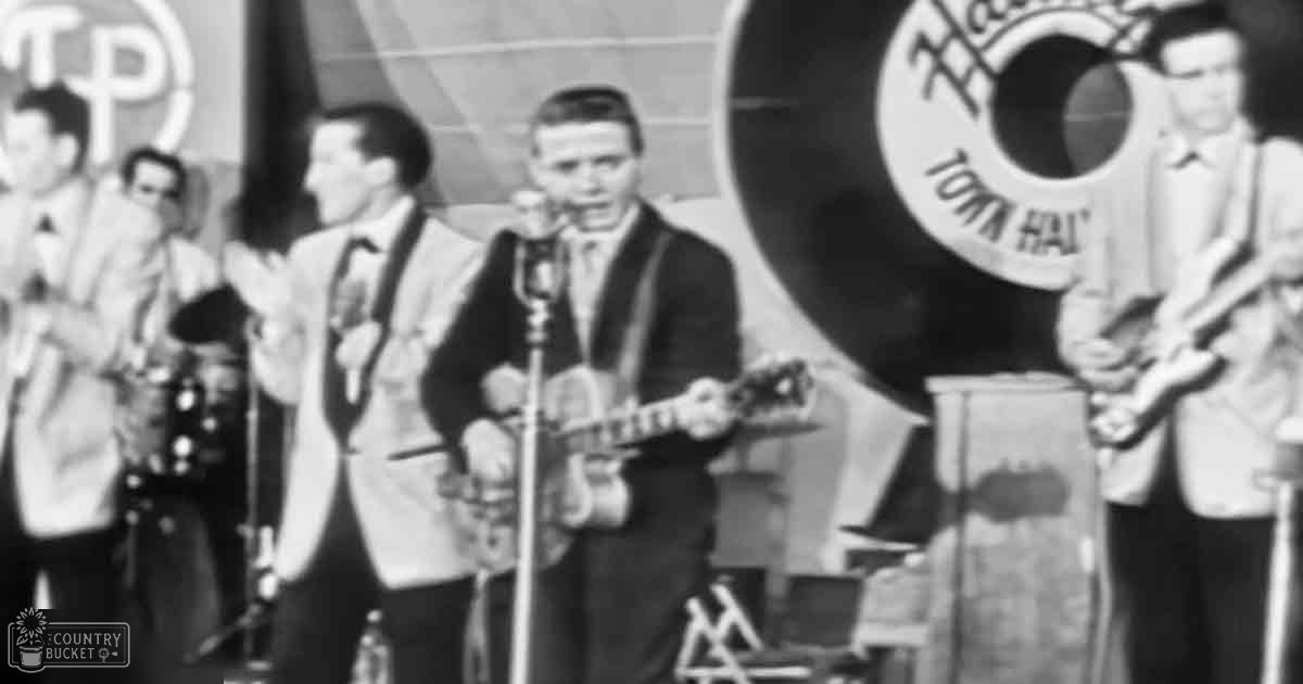 Musical performance with multiple adult male singers playing guitars, mandolins, or ukuleles on a stage set against a 'Town Hall' sign under professional indoor lighting.