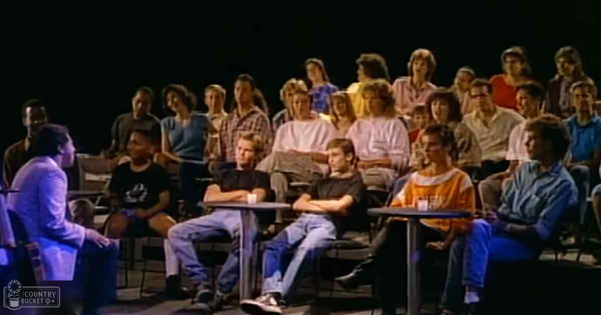 Indoor studio setting with dark lighting and audience members seated facing forward during a talk show or panel discussion.
