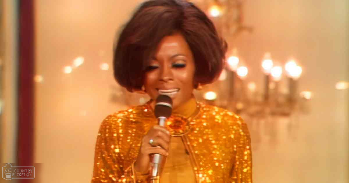 An adult female performer wearing a gold sequined 1960s-style outfit, holding a microphone in an indoor setting with warm ambient lighting and blurred lights suggesting a theater or concert hall atmosphere.