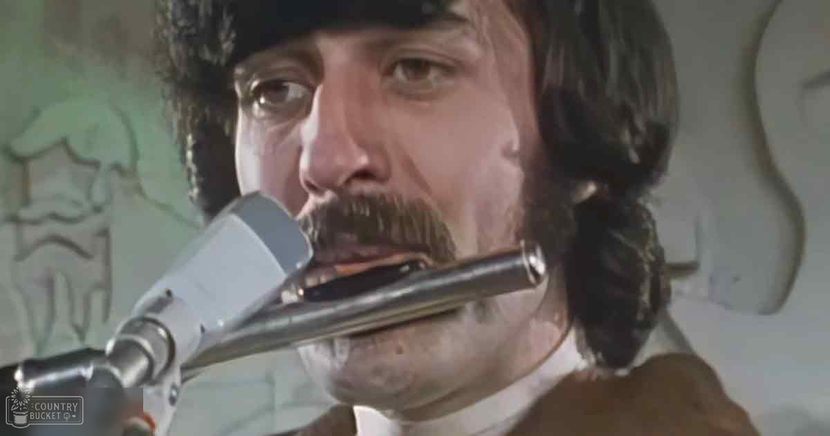 Adult male singer wearing a white, light cotton or linen mid-20th century-style garment with a high collar in an indoor recording studio setting, microphone prominently featured.