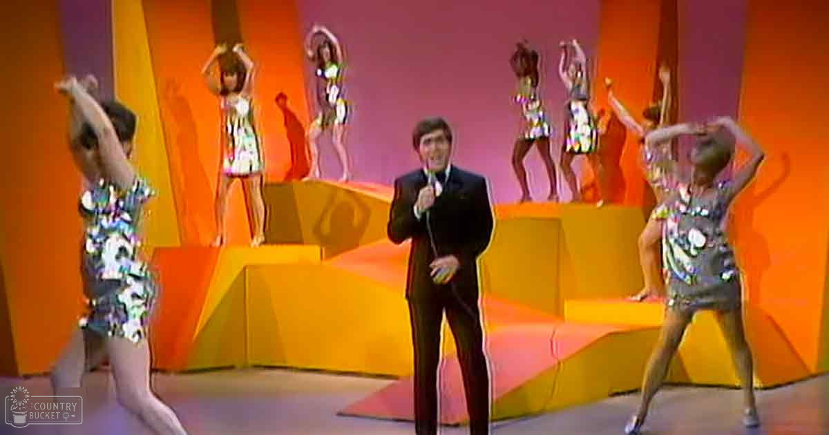 A middle-aged adult male singer holding a microphone performs with young female dancers in shiny, sequined outfits against an indoor stage set with vibrant orange and pink lighting.