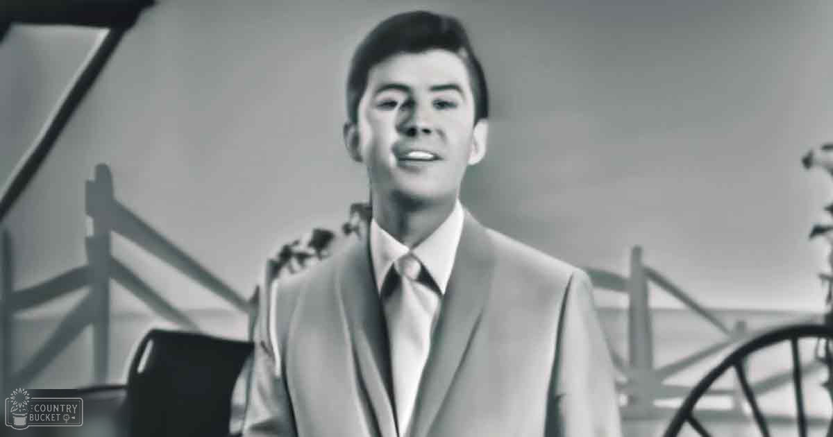 A young adult male wearing a tailored suit jacket with a tie stands indoors; the setting includes a chair and a staircase, suggesting a home environment.