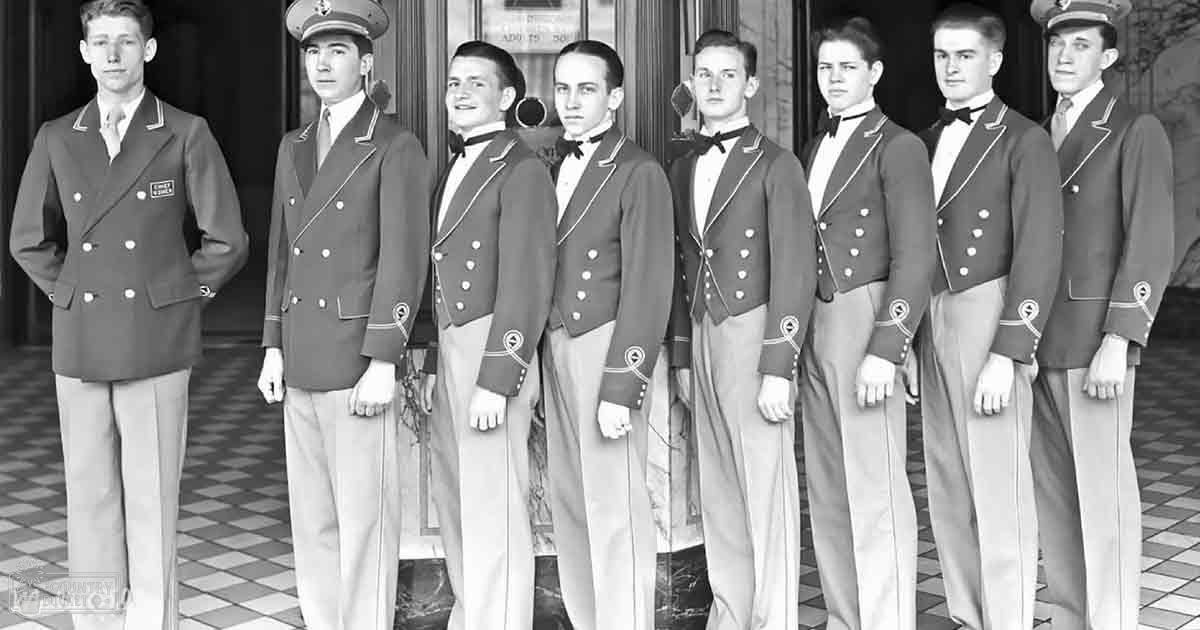 A group of young adult males in double-breasted blazers standing outside a building with tiled flooring, wearing light-colored trousers and decorative elements on their sleeves under natural daylight.