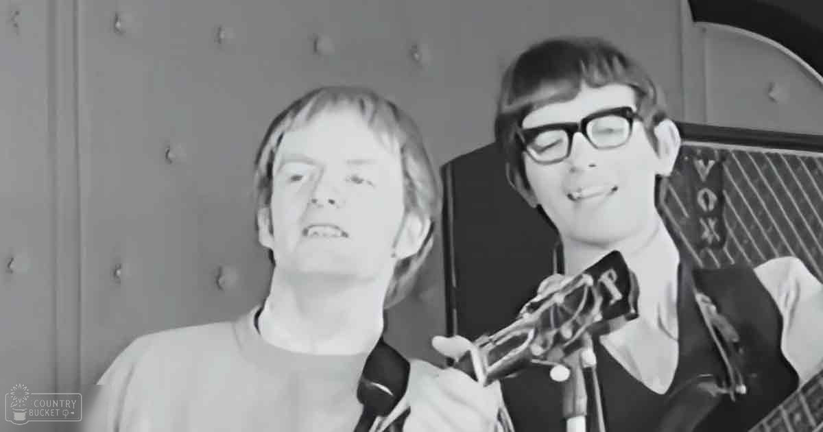 Young adult male musical duo in an indoor music practice or performance setting, wearing casual attire with a microphone stand and guitar visible in the frame.