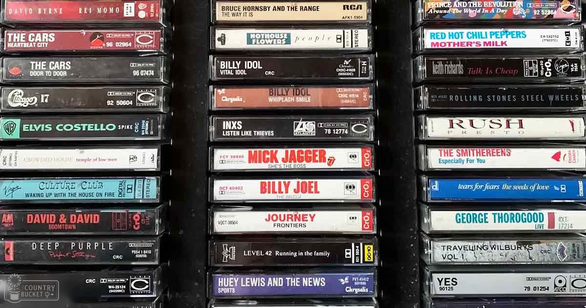 Stacks of cassette tapes from Billy Idol, Journey, Deep Purple, Elvis Costello in an indoor storage area with artificial lighting and various album cover colors.