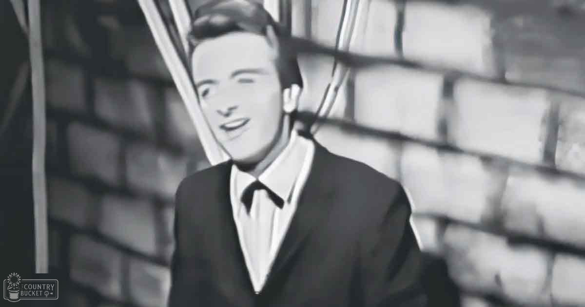 A young adult male in a dark suit jacket and trousers stands against a brick wall with scaffolding visible behind him, wearing a white shirt and black tie; text reading 'Madly Odd' is seen in the bottom left corner. The scene takes place outdoors during daytime.