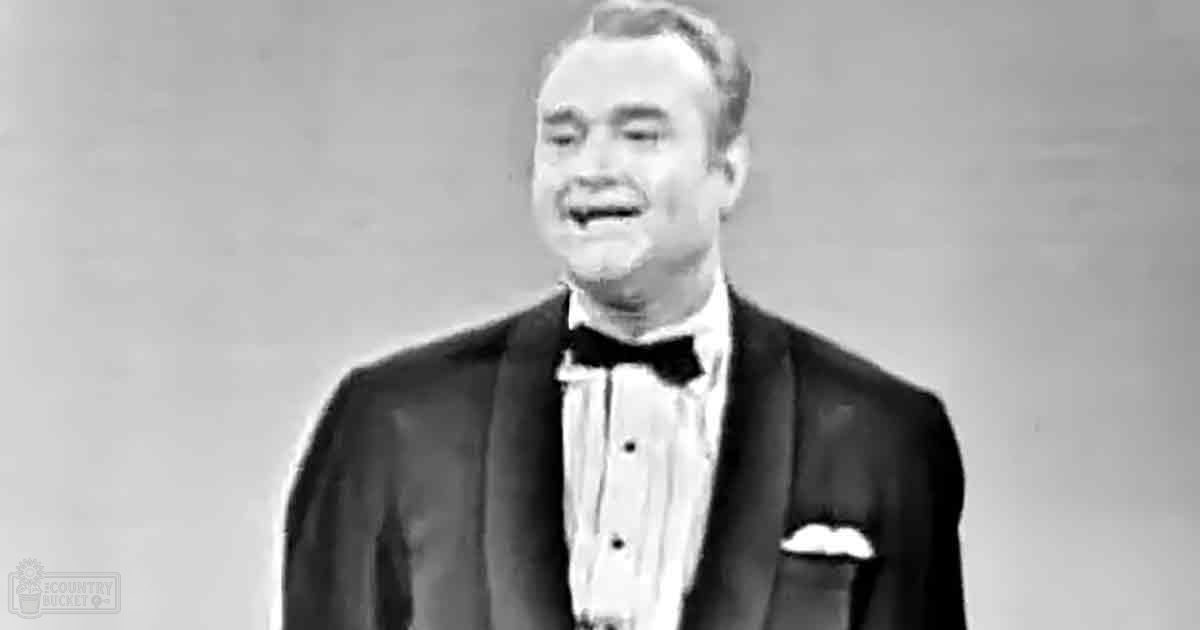 Middle-aged adult male wearing a well-fitted tuxedo with a bow tie, standing indoors against a plain light-colored wall.