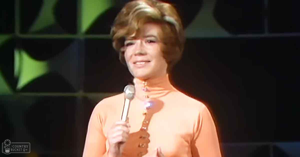 An adult female singer in a peach-colored 1960s-style dress holds a microphone, performing on stage with bright lighting and green geometric decor.