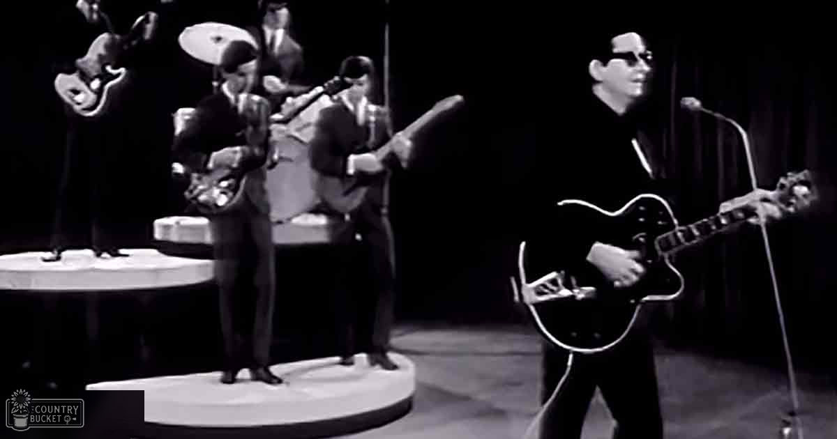 A mid-20th century musical performance scene with five adult male singers in dark suits, positioned on a stage setup with microphones and musical instruments under artificial studio lighting.