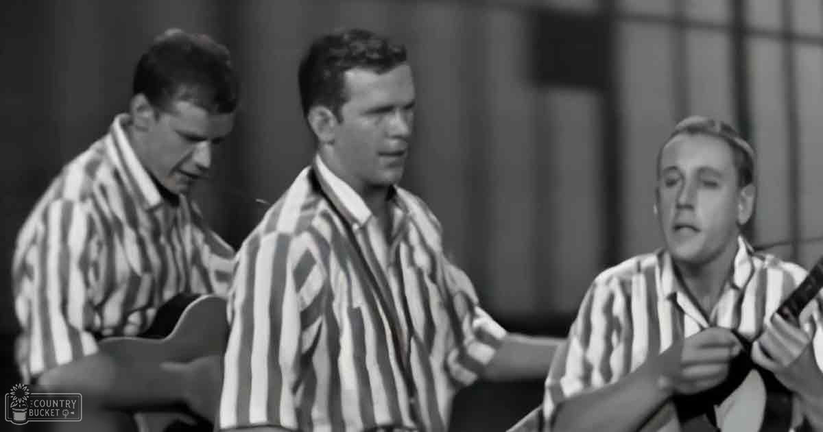 A group performance scene with three young adult males wearing striped shirts, playing guitars on a stage set for a musical production or prison-themed performance.