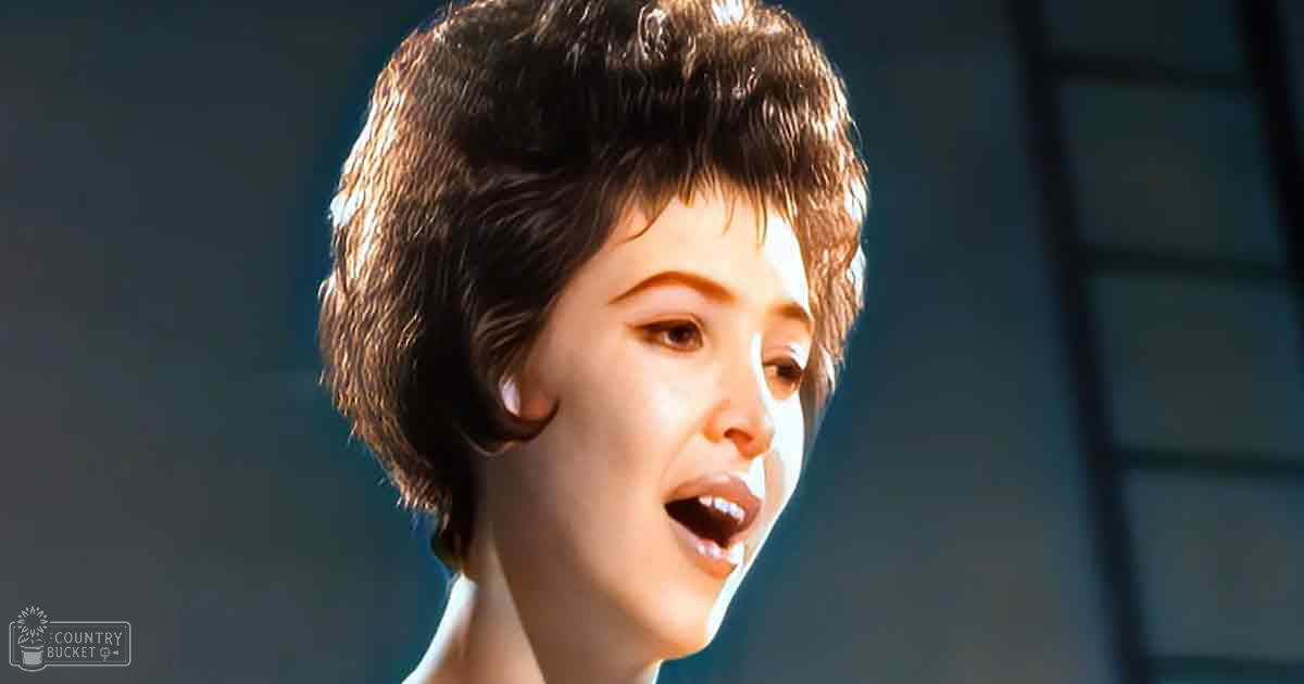 A young adult female in a white garment with high neckline and short sleeves, set against a dark indoor background suggesting evening time.