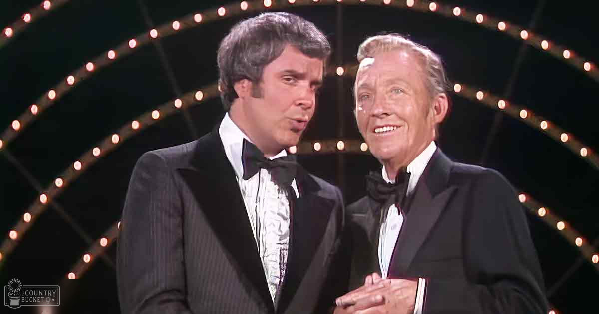 Two men in formal tuxedos stand on a stage, wearing classic black tuxedos with white dress shirts and black bow ties against an indoor venue backdrop.