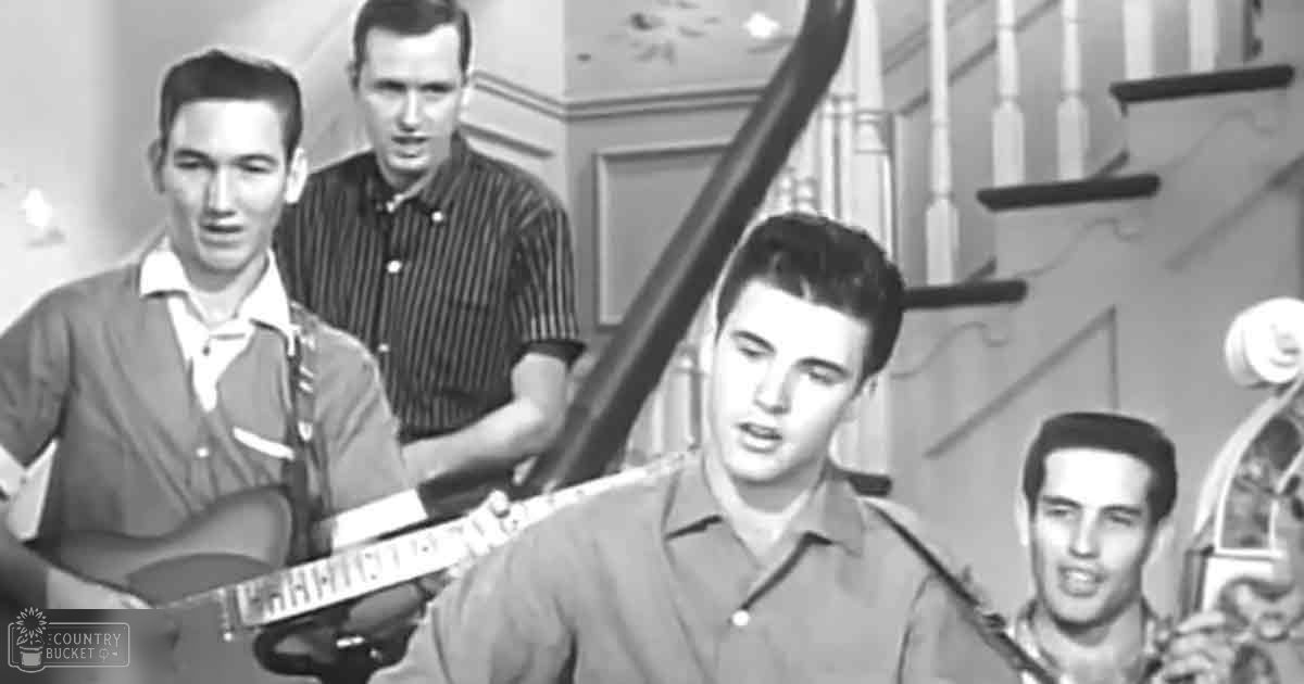 A young adult male is seen indoors near a staircase with wooden railings, wearing a light-colored collared shirt. An adult male in the background appears to be speaking while also wearing a vertically striped button-up shirt.