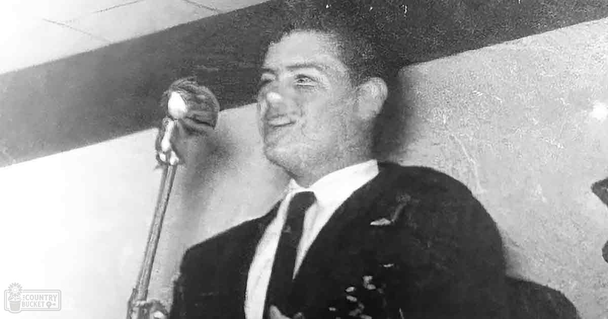 A young adult male in a dark suit jacket, white shirt, and tie stands indoors against plain walls with a microphone stand visible behind him.