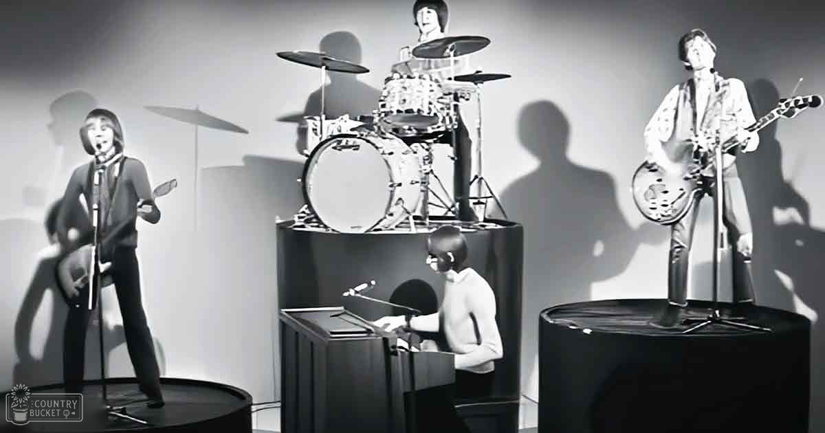 A wax museum display with a band from the 1960s era with three adult males in suits standing on black cylindrical platforms, each with microphones and guitars; plain white background, even bright lighting typical of indoor displays;