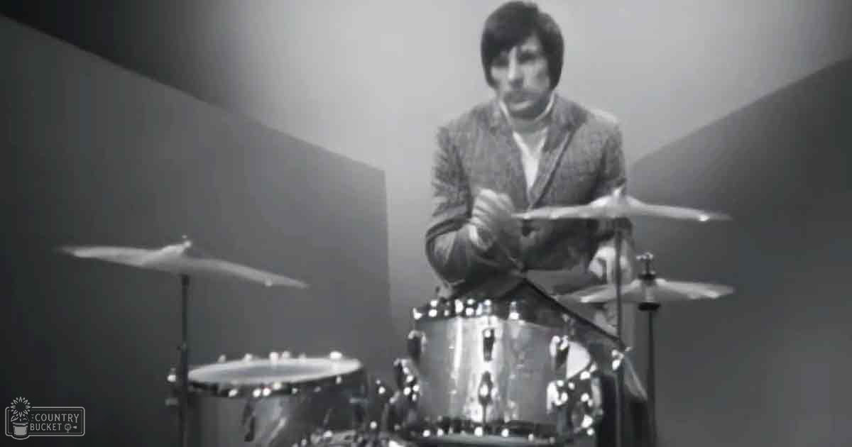 A young adult male drummer in a textured suit jacket performing indoors, surrounded by musical instruments under overhead lighting.